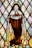A stained glass artwork depicting a religious figure dressed in a brown robe and white cloak, holding a banner that reads 'Follow Him.' The figure has a halo and three red roses on the robe. The background consists of diamond-shaped pieces of glass in subtle colors.