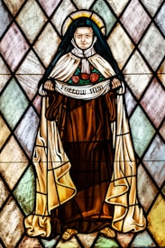 A stained glass artwork depicting a religious figure dressed in a brown robe and white cloak, holding a banner that reads 'Follow Him.' The figure has a halo and three red roses on the robe. The background consists of diamond-shaped pieces of glass in subtle colors.