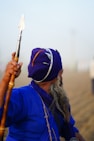 A person wearing a deep blue traditional outfit and matching turban, adorned with a metal accessory. They are holding a long wooden staff with a metal spearhead while facing away from the camera. The background is a foggy or misty outdoor setting.
