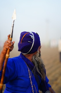 A person wearing a deep blue traditional outfit and matching turban, adorned with a metal accessory. They are holding a long wooden staff with a metal spearhead while facing away from the camera. The background is a foggy or misty outdoor setting.
