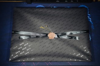 A rectangular object is wrapped in branded black paper featuring the word 'GRID' in a repeating pattern. A black ribbon with 'Frame Studio' text is tied around it, sealed with a wax-like stamp in the center. The package sits on a blue and dark patterned surface.