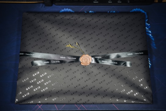 A rectangular object is wrapped in branded black paper featuring the word 'GRID' in a repeating pattern. A black ribbon with 'Frame Studio' text is tied around it, sealed with a wax-like stamp in the center. The package sits on a blue and dark patterned surface.