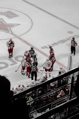 A group of hockey players in red and white uniforms gather near the center of an ice rink. Several referees in black and white striped jerseys are present among the players. Spectators can be seen seated in the stands, watching the game from behind a glass barrier. The ice surface is marked with lines and a large star logo.