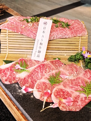 Slices of marbled Wagyu beef are elegantly arranged on a wooden mat, garnished with fresh greens and pink flowers. A small wooden sign written in Japanese and English identifies the meat as A5 grade Wagyu. The setup is enhanced by a dark stone surface, providing a contrast to the vibrant colors of the beef.