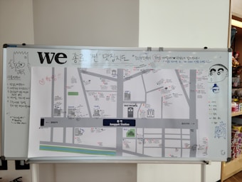 A large whiteboard features a map with various locations marked in the Jonggak Station area. The map includes handwritten notes, drawings of cartoon characters, and a variety of colored markers. The board has a playful and informal atmosphere with doodles and exclamations.