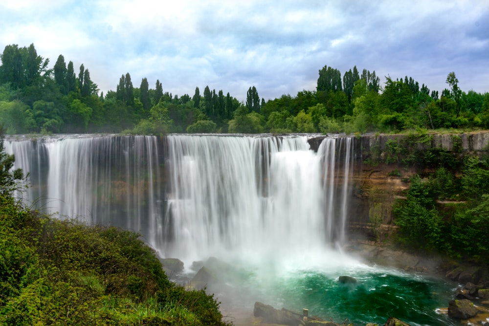 waterfalls near green trees under white clouds during daytime