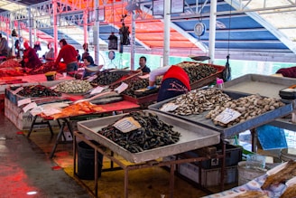 Stalls in a bustling seafood market display a wide variety of fresh shellfish and fish. Several trays are filled with mussels, clams, and other seafood. People are busy organizing and selling their goods, while colorful signs indicate prices. The market space is covered, with bright colored awnings casting vibrant hues over the scene.