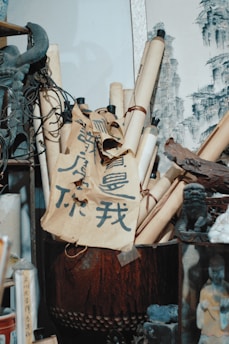 A collection of scrolls and calligraphy papers are neatly arranged in a pot. The scrolls are tied with black cords, and there is bold, stylized writing in dark ink on the beige paper. Traditional art is visible in the background, featuring monochromatic sketches of cliffs. Surrounding the scrolls are various artifacts, including a statue of a mythical creature and other decor items, contributing to an atmosphere reminiscent of an antique shop or art studio.