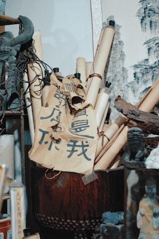 A collection of scrolls and calligraphy papers are neatly arranged in a pot. The scrolls are tied with black cords, and there is bold, stylized writing in dark ink on the beige paper. Traditional art is visible in the background, featuring monochromatic sketches of cliffs. Surrounding the scrolls are various artifacts, including a statue of a mythical creature and other decor items, contributing to an atmosphere reminiscent of an antique shop or art studio.