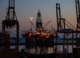 An offshore oil platform is illuminated against the evening sky. Large cranes surround the structure, and the sea is visible in the background. The lighting suggests it is dusk, with the horizon showing a blend of dark blue and orange hues.