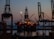 An offshore oil platform is illuminated against the evening sky. Large cranes surround the structure, and the sea is visible in the background. The lighting suggests it is dusk, with the horizon showing a blend of dark blue and orange hues.
