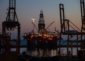An offshore oil platform is illuminated against the evening sky. Large cranes surround the structure, and the sea is visible in the background. The lighting suggests it is dusk, with the horizon showing a blend of dark blue and orange hues.