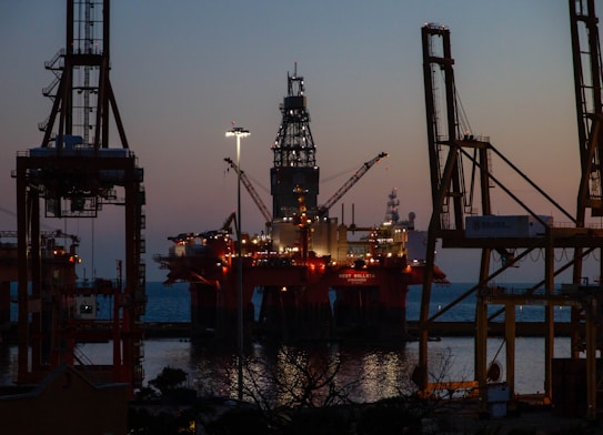 An offshore oil platform is illuminated against the evening sky. Large cranes surround the structure, and the sea is visible in the background. The lighting suggests it is dusk, with the horizon showing a blend of dark blue and orange hues.