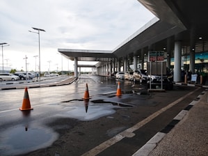 A modern transportation hub with a sleek architectural design featuring a covered walkway and multiple vehicles parked along the road. Orange traffic cones are placed on the wet road surface, and puddles from recent rain are visible. The area is well-lit with street lamps.