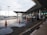 A modern transportation hub with a sleek architectural design featuring a covered walkway and multiple vehicles parked along the road. Orange traffic cones are placed on the wet road surface, and puddles from recent rain are visible. The area is well-lit with street lamps.
