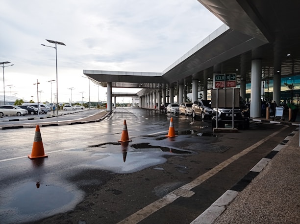 A modern transportation hub with a sleek architectural design featuring a covered walkway and multiple vehicles parked along the road. Orange traffic cones are placed on the wet road surface, and puddles from recent rain are visible. The area is well-lit with street lamps.