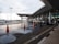 A modern transportation hub with a sleek architectural design featuring a covered walkway and multiple vehicles parked along the road. Orange traffic cones are placed on the wet road surface, and puddles from recent rain are visible. The area is well-lit with street lamps.