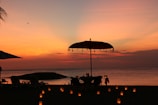 A tranquil beach scene at sunset with a large umbrella, a table and chairs set for dining near the water. Lit candles are arranged on the sand, creating a warm and inviting atmosphere. The ocean is calm, and the sky is painted in vibrant shades of orange, pink, and yellow.