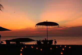 A tranquil beach scene at sunset with a large umbrella, a table and chairs set for dining near the water. Lit candles are arranged on the sand, creating a warm and inviting atmosphere. The ocean is calm, and the sky is painted in vibrant shades of orange, pink, and yellow.