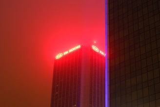 A tall building with the top section illuminated by bright red and yellow lights displaying the words 'KIA MOTORS'. The background is a hazy sky with a reddish tint, possibly from artificial lighting or pollution. Another building partially obscures the view to the right, with a vertical line of blue light running along its edge.