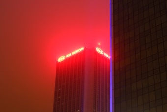 A tall building with the top section illuminated by bright red and yellow lights displaying the words 'KIA MOTORS'. The background is a hazy sky with a reddish tint, possibly from artificial lighting or pollution. Another building partially obscures the view to the right, with a vertical line of blue light running along its edge.