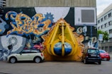 A vibrant graffiti mural covers a large wall, depicting an industrial scene with gears and vibrant abstract designs. A large, three-dimensional yellow structure resembling a construction claw is integrated into the mural, giving an illusion of bursting through the wall. Three cars are parked in front, adding a sense of urban environment.