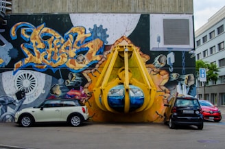A vibrant graffiti mural covers a large wall, depicting an industrial scene with gears and vibrant abstract designs. A large, three-dimensional yellow structure resembling a construction claw is integrated into the mural, giving an illusion of bursting through the wall. Three cars are parked in front, adding a sense of urban environment.
