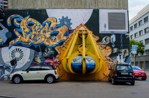 A vibrant graffiti mural covers a large wall, depicting an industrial scene with gears and vibrant abstract designs. A large, three-dimensional yellow structure resembling a construction claw is integrated into the mural, giving an illusion of bursting through the wall. Three cars are parked in front, adding a sense of urban environment.