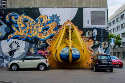 A vibrant graffiti mural covers a large wall, depicting an industrial scene with gears and vibrant abstract designs. A large, three-dimensional yellow structure resembling a construction claw is integrated into the mural, giving an illusion of bursting through the wall. Three cars are parked in front, adding a sense of urban environment.