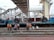 A group of people are gathered around train tracks in what appears to be a railway station. Large coils of metal are loaded onto a blue flatbed train car. Overhead, there is a pedestrian bridge connecting different platforms, and the structure of the station is visible in the background.