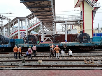 A group of people are gathered around train tracks in what appears to be a railway station. Large coils of metal are loaded onto a blue flatbed train car. Overhead, there is a pedestrian bridge connecting different platforms, and the structure of the station is visible in the background.