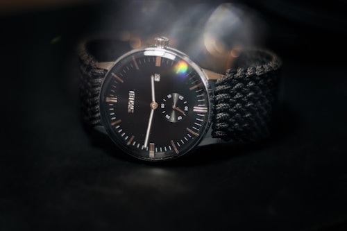 A close-up photograph of a wristwatch with a classic design, featuring a black dial with hour markers and a smaller sub-dial for seconds. The watch has a stainless steel case and a textured black strap. The background is dark, accentuating the watch's details with a slight lens flare effect near the top right of the watch face.