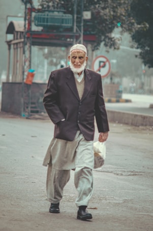 An elderly man is walking on a street. He is dressed in traditional clothing with a dark blazer and a light gray shalwar kameez. He wears a white cap and is holding a white bag. The background shows an urban setting with a bus stop, a tree, and various street elements.