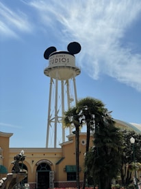 A large water tower with Mickey Mouse ears is prominently visible against a clear blue sky. The lower section reveals a building with soft yellow walls and green awnings. Palm trees and other greenery surround the area, adding a touch of natural scenery.