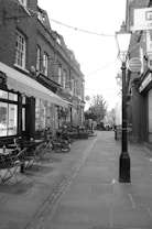 A narrow cobblestone alleyway flanked by traditional brick buildings. Small tables and chairs are set outside a caf&eacute;, suggesting a quaint, inviting atmosphere. Potted plants and hanging lights line the alley, adding a touch of greenery and charm. The scene appears serene and picturesque.