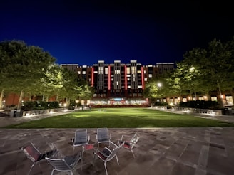 An urban courtyard at night featuring a modern multi-story building illuminated with lights. Trees line the courtyard, creating a symmetrical path leading to the building. Several empty chairs are scattered on the stone patio area in the foreground, and the grass in the middle is neatly maintained.