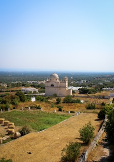 A serene landscape featuring a historic church with a dome and two bell towers surrounded by lush green fields and trees. The structure is nestled in a rural setting with scattered homes and farm fields leading to a vast horizon under a bright blue sky.