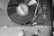 A black and white image of a vinyl record player with a spinning record. The turntable is positioned in the upper part of the image, with the tonearm placed on the record. The control knobs and buttons are on the right side of the player. The view also shows a pair of blurred feet wearing socks at the bottom of the frame.