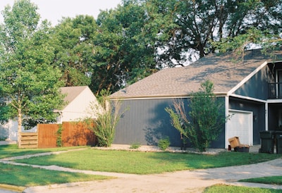 A suburban house with a dark exterior and a sloped roof is surrounded by lush greenery. The neatly manicured lawn and a few tall trees contribute to a serene, residential atmosphere. A wooden fence separates the house from the adjacent property. The driveway leads to a closed garage door and some trash bins are placed nearby.