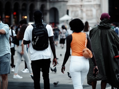 A busy urban setting with people walking in various directions. The central focus is on two individuals walking together; one wearing a black cap, white shirt, and black backpack, and the other wearing a bright orange top and white pants with an orange purse. The scene includes other pedestrians and pigeons scattered on the ground. Architectural details are visible in the background.