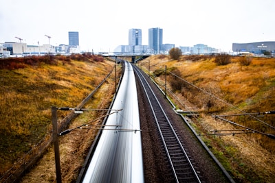 A high-speed train travels through a landscape with grassy embankments flanking both sides of the railway line. The train is blurred due to its speed, emphasizing motion. In the background, modern high-rise buildings and industrial structures are visible under a clear sky.