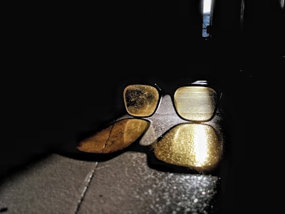A pair of sunglasses lies on a surface, reflecting sunlight that creates a warm, golden glow. The light casts long shadows and highlights the texture and details on the lenses, contrasting sharply with the dark surrounding environment.