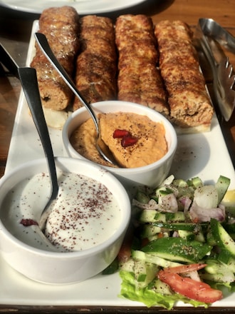 A platter of grilled kebabs served with two types of dipping sauces and a fresh vegetable salad. The kebabs are arranged in a row, appearing juicy and well-cooked. The sauces include a creamy white sauce with sprinkled seasoning and a reddish-orange hummus garnished with pomegranate seeds. The salad consists of sliced cucumbers, tomatoes, onions, and lettuce, seasoned with herbs.