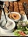 A platter of grilled kebabs served with two types of dipping sauces and a fresh vegetable salad. The kebabs are arranged in a row, appearing juicy and well-cooked. The sauces include a creamy white sauce with sprinkled seasoning and a reddish-orange hummus garnished with pomegranate seeds. The salad consists of sliced cucumbers, tomatoes, onions, and lettuce, seasoned with herbs.