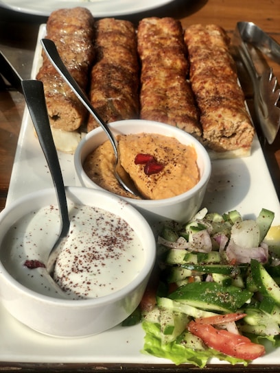 A platter of grilled kebabs served with two types of dipping sauces and a fresh vegetable salad. The kebabs are arranged in a row, appearing juicy and well-cooked. The sauces include a creamy white sauce with sprinkled seasoning and a reddish-orange hummus garnished with pomegranate seeds. The salad consists of sliced cucumbers, tomatoes, onions, and lettuce, seasoned with herbs.