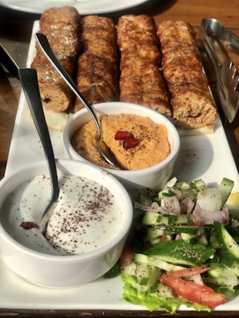 A platter of grilled kebabs served with two types of dipping sauces and a fresh vegetable salad. The kebabs are arranged in a row, appearing juicy and well-cooked. The sauces include a creamy white sauce with sprinkled seasoning and a reddish-orange hummus garnished with pomegranate seeds. The salad consists of sliced cucumbers, tomatoes, onions, and lettuce, seasoned with herbs.