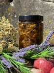 A jar filled with honeycomb sits in natural lighting against a rustic stone background. Surrounding the jar are sprigs of dried flowers and fresh lavender, alongside a couple of red apples.