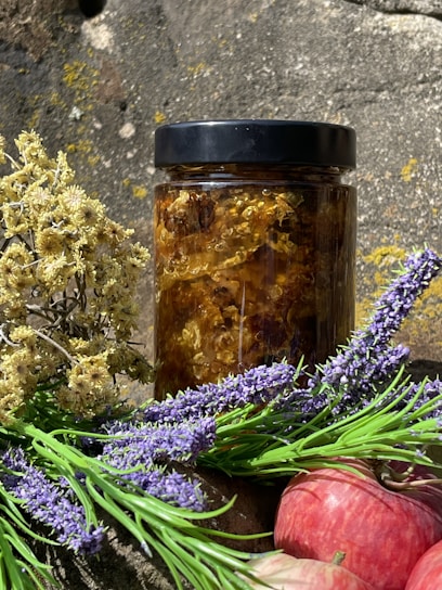 A jar filled with honeycomb sits in natural lighting against a rustic stone background. Surrounding the jar are sprigs of dried flowers and fresh lavender, alongside a couple of red apples.