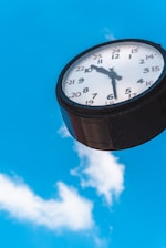 A round clock with a white face and black numbers, including a 24-hour format, is mounted against a bright blue sky with scattered white clouds. The clock's hands indicate a time of around 10:10.