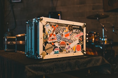 A large metal flight case covered in various colorful stickers sits on a table. The background reveals a drum set partially visible in a dimly lit room with a rustic wall, creating an ambiance often found in music venues.